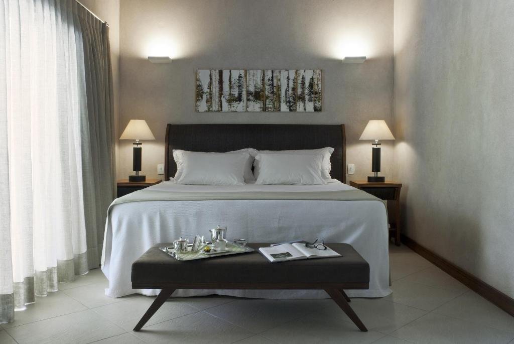 Abayomi Hotel. Ilustra o post sobre hotéis em Ilhabela. O quarto tem paredes claras e um piso escuro.
Há uma cama grande com uma cabeceira escura, lençóis brancos, dois travesseiros e duas almofadas decorativas.
Acima da cama, há uma peça de arte retangular que parece representar árvores sem folhas.
De cada lado da cama, há mesinhas de cabeceira escuras com abajures modernos que estão acesos.
Uma cortina branca cobre uma janela à esquerda da imagem.
Na frente da cama, há um banco escuro com pernas angulares. Em cima do banco, há uma bandeja com duas taças de vinho, uma garrafa e um livro aberto ao lado dela.