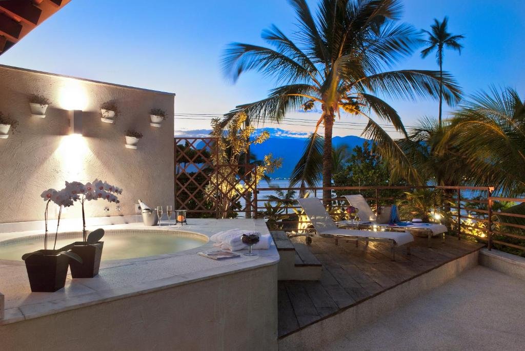 Abayomi Hotel. A imagem mostra uma varanda ao ar livre com vista para um cenário tropical.
Há uma jacuzzi embutida na varanda, com água calma e iluminação suave.
Uma planta em vaso com flores brancas está próxima à jacuzzi.
O piso da varanda é de madeira, e há uma cadeira confortável e uma mesa pequena colocadas estrategicamente para apreciar a vista.
A varanda é cercada por um corrimão de madeira, adornado com luzes decorativas que proporcionam um ambiente acolhedor.
Palmeiras altas e exuberantes estão visíveis ao fundo, destacando-se contra o céu azul claro do entardecer ou amanhecer.
As montanhas distantes são visíveis no horizonte, adicionando profundidade e beleza natural à cena.