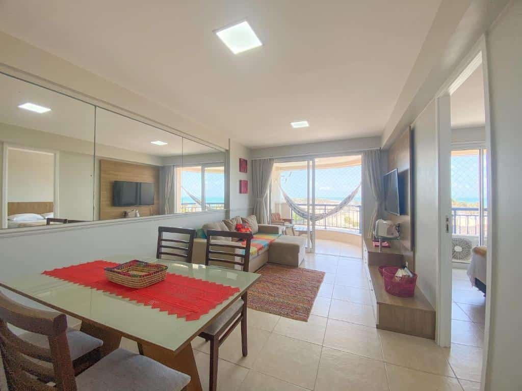 Sala de estar do AllMar Flats - Apartamentos frente mar - Beach Village. Em primeiro plano está uma mesa de quatro lugares, ao lado de uma porta aberta. Ao fundo está a sala de estar com um sofá de frente para uma tv. Ao lado da sala está uma varanda com rede.