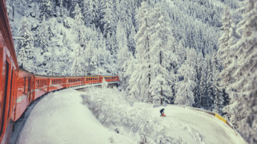 Um trem vermelho se move por uma floresta coberta de neve, passando por um esquiador em uma encosta. A cena é envolta em cenário de inverno.