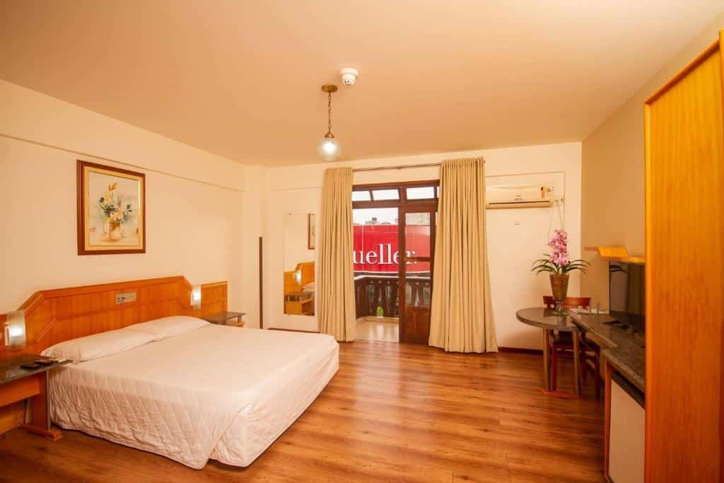 Quarto do Alven Palace Hotel em Joinville com uma cama de casal à esquerda e uma mesa com frigobar e televisão à direita. Entre esses objetos há uma sacada com vista para o shopping mueller