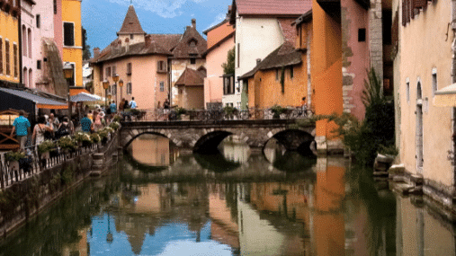 Um canal pitoresco ladeado por edifícios coloridos em Annecy e uma ponte de pedra refletida na água, sob um céu parcialmente nublado. Representa passeios na Suíça.