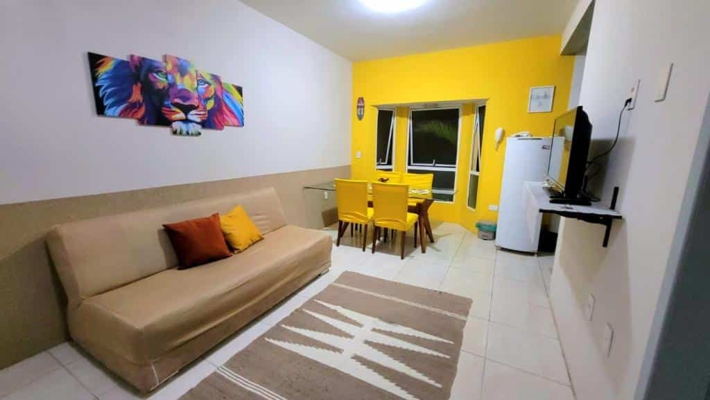 Sala de estar do Apartamento Caminho do Mar. Um sofá e um tapete do lado esquerdo, de frente uma televisão. No fundo, do lado direito uma geladeira e um lixo, do lado esquerdo uma mesa de vidro com cadeiras e no fundo janelas.