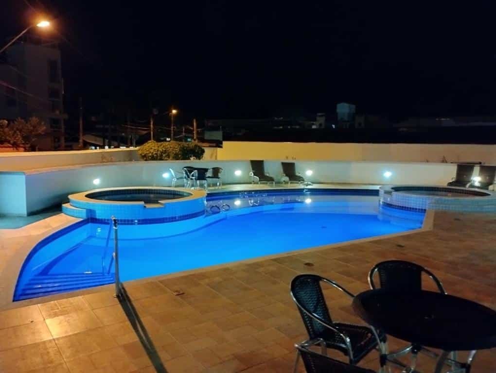 Área externa do Apto com ar-cond e piscina. Uma piscina de adulto e uma infantil de cada lado. Ao redor cadeiras de tomar sol, cadeiras e mesas.