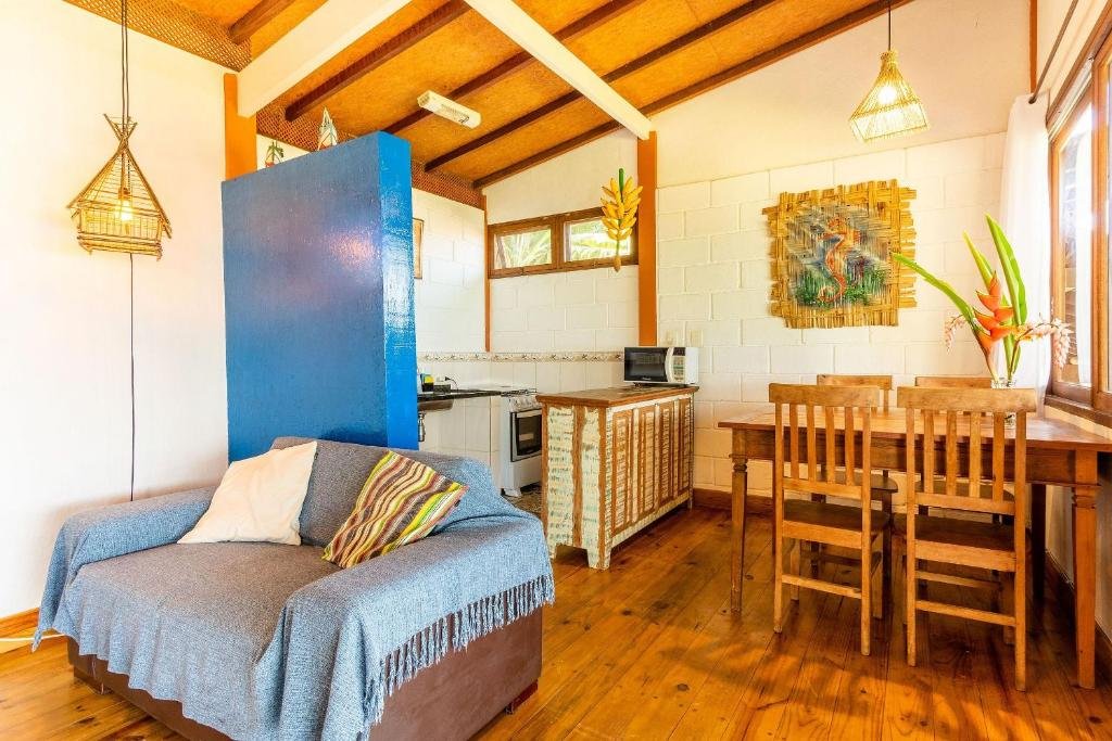 Visão geral do Bangalô Ilhabela. Ao lado esquerdo fica a sala de estar, onde está um sofá com manta e almofadas. Ao lado direito está uma mesa de madeira com quatro cadeiras e uma janela. Uma parede separa a sala da cozinha, onde é possível ver um fogão, um micro-ondas e bancadas na pia.