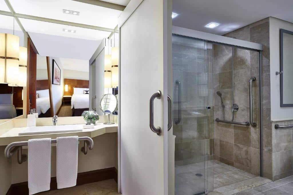 Imagem do banheiro. No lado esquerdo uma pia grande e baixa com barras. No lado direito o box de vidro com barras.