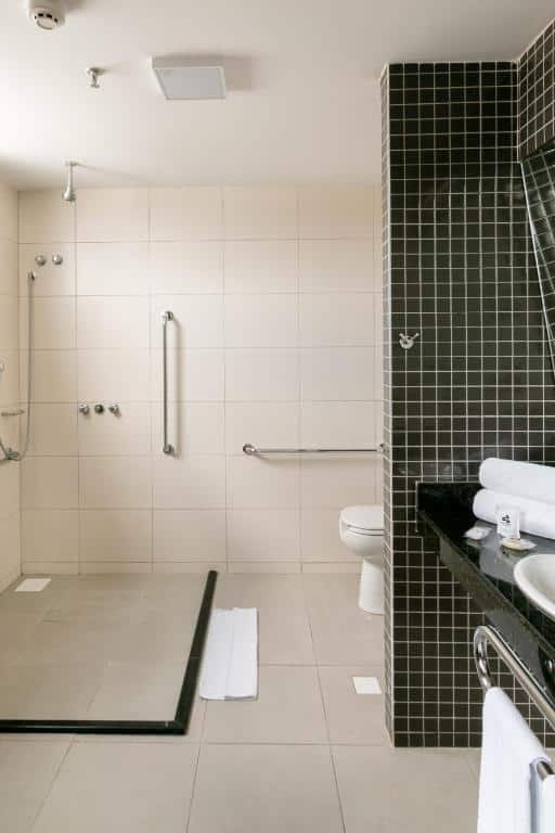 Banheiro do WA Hotel Caruaru. O banheiro é amplo, o chuveiro sai do teto e não tem paredes para dividir o box. Na parede ao fundo tem duas barras de apoio, uma privada no lado direito e na frente é possível ver uma parte da pia com mais uma barra de apoio. Imagem para ilustrar o post hotéis em Caruaru.