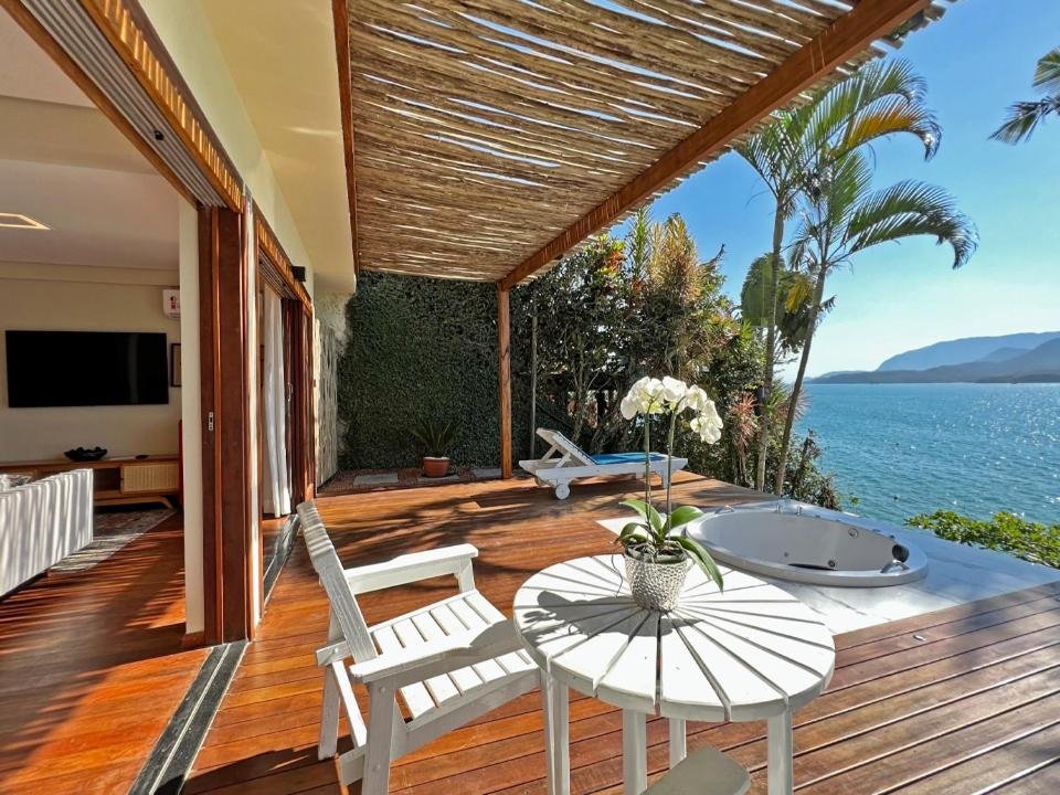 Barra do Piuva Porto Hotel. O terraço é coberto por um telhado de palha, proporcionando sombra e um ambiente tropical.
Há duas cadeiras brancas e uma mesa branca redonda no centro do terraço, com um vaso de flores brancas em cima.
Uma banheira de hidromassagem está visível à direita, oferecendo uma vista panorâmica do oceano.
O piso do terraço é feito de tábuas de madeira polidas e bem mantidas.
À esquerda, há uma parede verde que parece ser coberta por plantas trepadeiras ou algum tipo de vegetação.
Através das folhas das palmeiras à direita, pode-se ver o mar azul claro e as montanhas ao fundo sob um céu ensolarado.