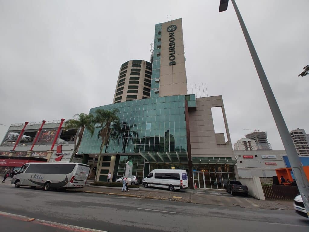 fachada do hotel bourbon joinville convention onde é possível enxergar a rua com alguns carros passando em frente, e duas torres no prédio, uma menor do que a outra, sendo a menor arredondada.