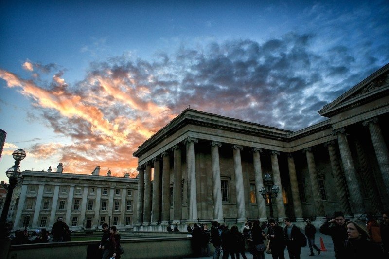 A imagem mostra um edifício neoclássico ao pôr do sol, com pessoas reunidas em primeiro plano e nuvens no céu. Esse é o British Museum, uma das atrações para adicionar ao roteiro para Londres.