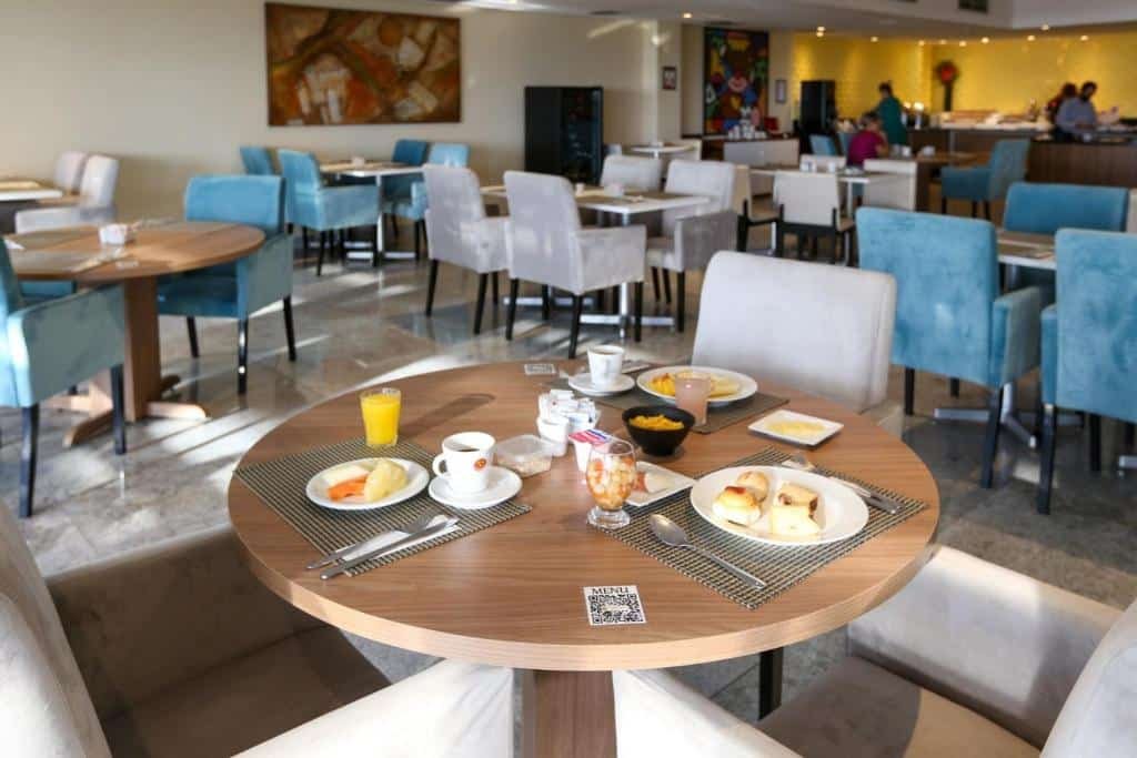Salão do café da manhã, com várias mesas e cadeiras. Na frente, uma mesa com o café da manhã, com café, suco, pães e frios.