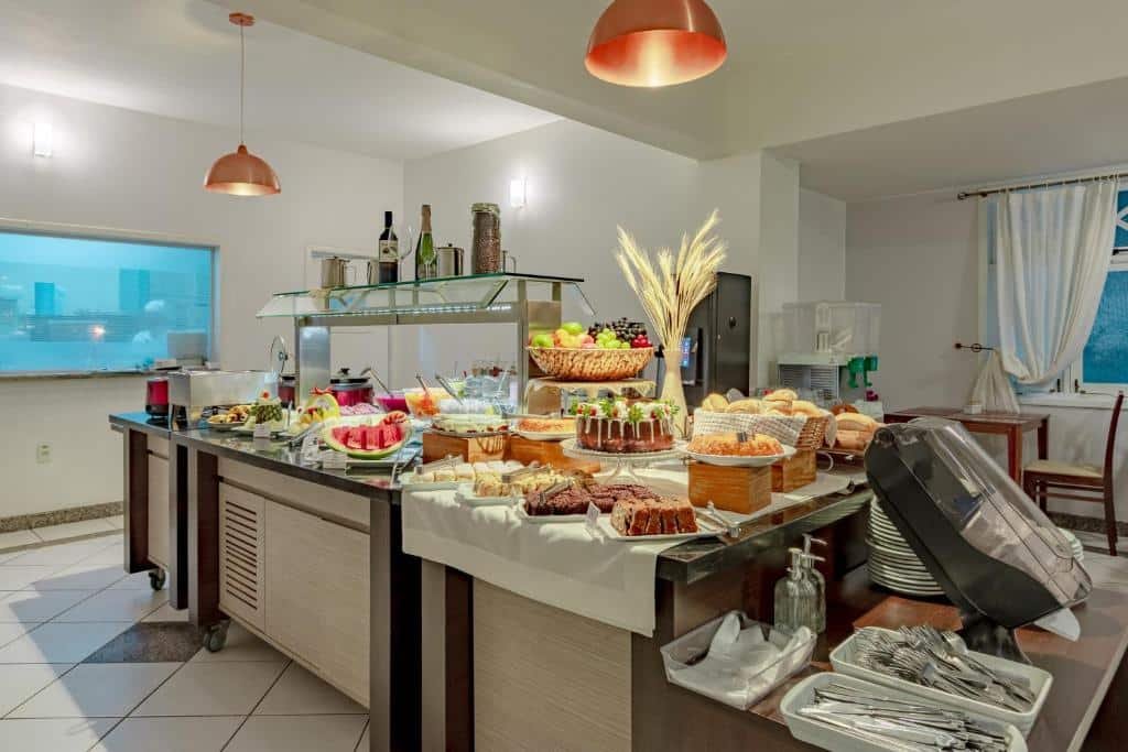 área de refeições do Bristol Sabrina Hotel com um balcão de alimentos no centro do espaço. Há várias frutas, bolos, pães e doces