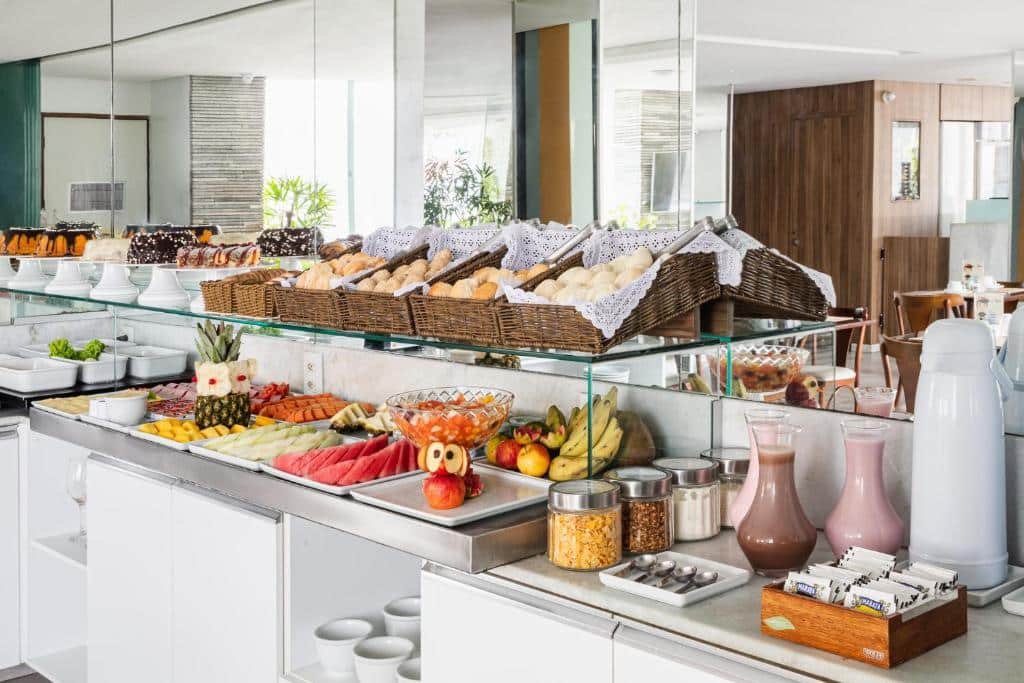Área de café da manhã do Citi Hotel Express Caruaru. Em cima de uma bancada estão diversas frutas, cereais, iogurtes, uma garrafa de café e sachês de chá. Em cima da bancada há uma prateleira de vidro com bolos, pães e salgados. Atrás na bancada há um espelho cobrindo a maior parte da parede.