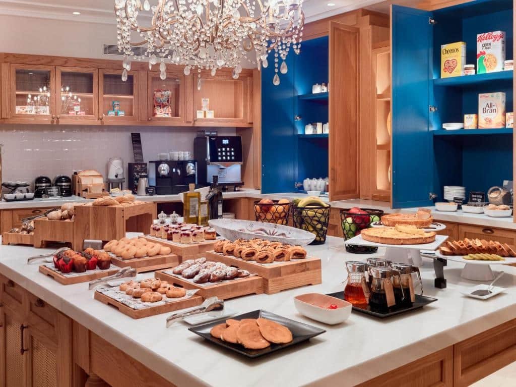 Mesa de café do H10 Casa de la Plata. Uma mesa quadrada no centro com um lustre, uma variedade de opções do café da manhã. Atrás armários no tom marrom e azul com copos, cafeteira e pratos. Foto para ilustrar post sobre Hotéis em Sevilha.