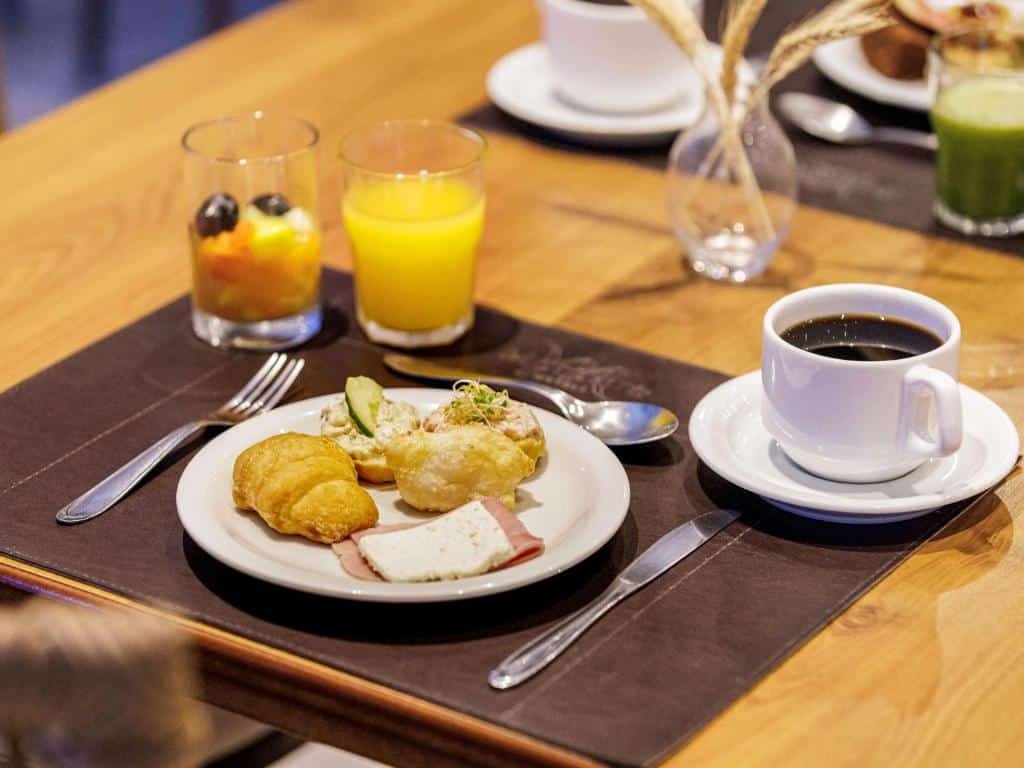 mesa com café da manhã no Mercure Joinville Prinz com um copo de suco de laranja, uma xícara branca com café, um copo de vidro com frutas picadas e um prato branco com quitutes.