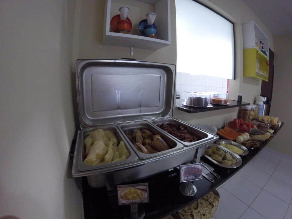 Bancada com o café da manhã servido na Pousada Sumaré. Em cima da bancada estão alimentos típicos da região, como a macaxeira e outros alimentos como frutas, pães, bolos e garrafas térmicas.