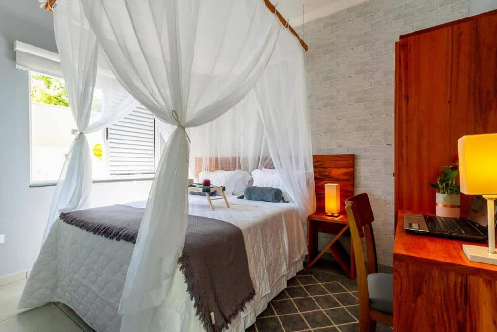 Quarto do Calamar Flats Paraty. Uma mesa de trabalho com um abajur e um computador do lado direito. Do lado esquerdo uma janela. No meio uma cama de casal com uma mesinha em cima e do lado uma mesa de apoio com um abajur. Foto para ilustrar post sobre airbnb em Paraty.