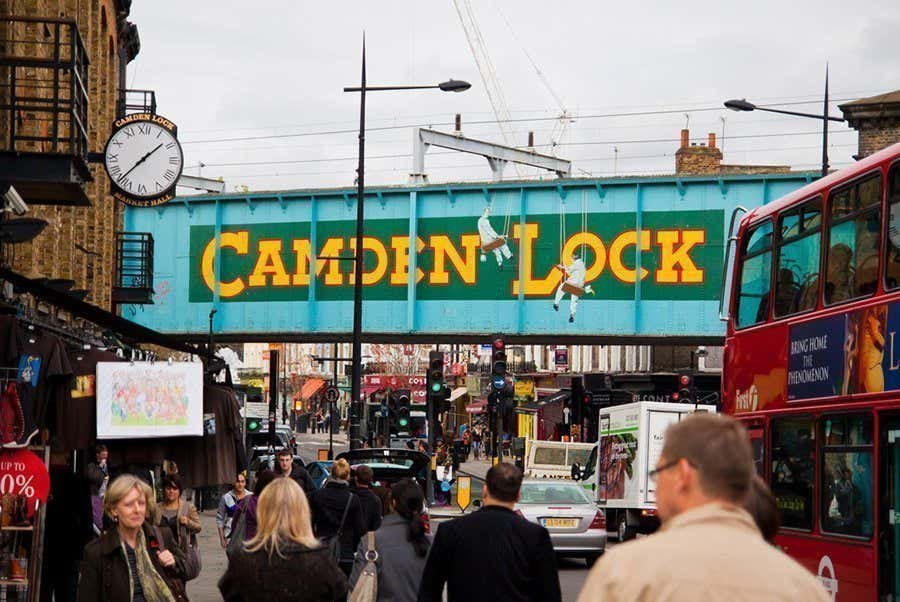 Cena de rua em Camden, Londres, com uma ponte azul acima exibindo um grande texto "Camden Lock". Pessoas e um ônibus vermelho são visíveis.
