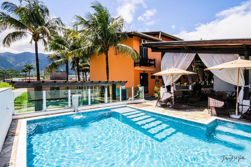 Parte da pousada CAMINHO DAS PRAIAS GUESTHOUSE que mostra uma piscina ao ar livre e do lado direito uma parte coberta com mesas e cadeiras. Ao fundo tem a construção laranja da pousada e algumas palmeiras grandes durante o dia. Imagem ilustrando post Pousadas pet friendly no Litoral Norte de São Paulo.