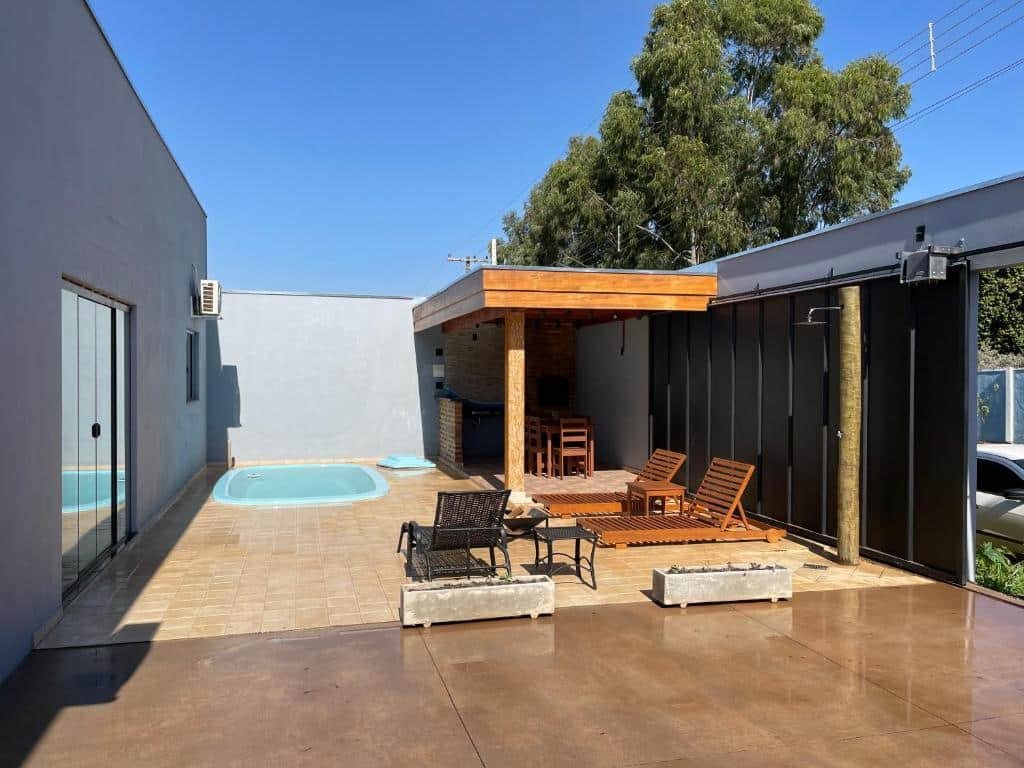 Parte do Cantinho Estrelado que mostra a parte externa da casa com um chão frio na garagem e depois um piso marrom. Na parte do piso tem algumas espreguiçadeiras, uma piscina e uma área coberta com uma churrasqueira e uma mesa de madeira. Imagem ilustrando post Airbnb em Brotas.