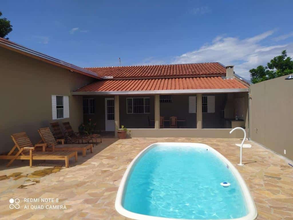Parte externa da "Casa acolhedora, bem aconchegante com piscina" com uma piscina, algumas espreguiçadeiras de madeira em volta e ao fundo o rancho coberto com uma mesa, uma churrasqueira, com portas e janelas aparecendo e um telhado de telha vermelha. Imagem ilustrando post Airbnb em Brotas.