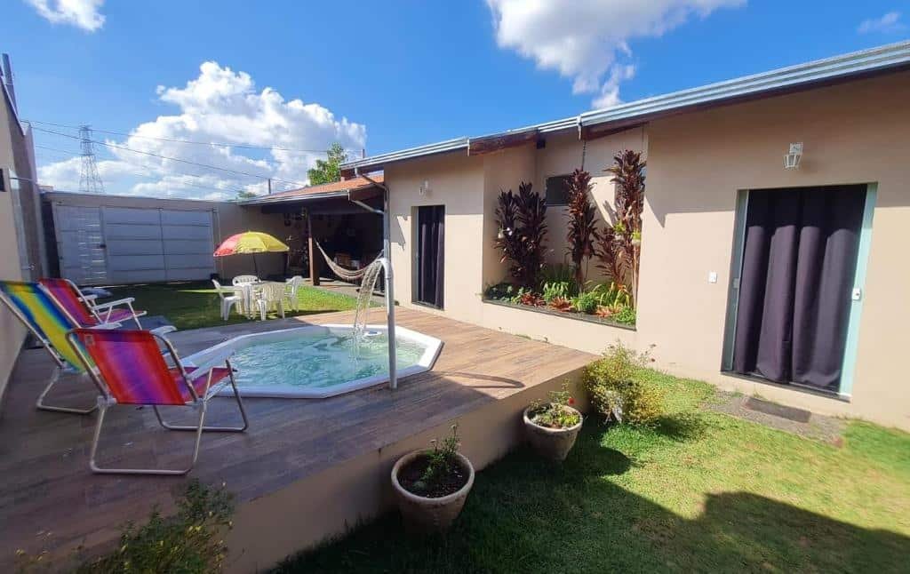 Parte externa da Casa c/ quartos separados + churrasqueira ampla com uma mini piscina com cascata e em volta algumas cadeiras de praia. Do lado direito da foto tem a construção do airbnb com duas portas com cortinas, gramado em volta e bem ao fundo uma mesa com um guarda-sol, um portão e uma parte coberta. Imagem ilustrando post Airbnb em Brotas.