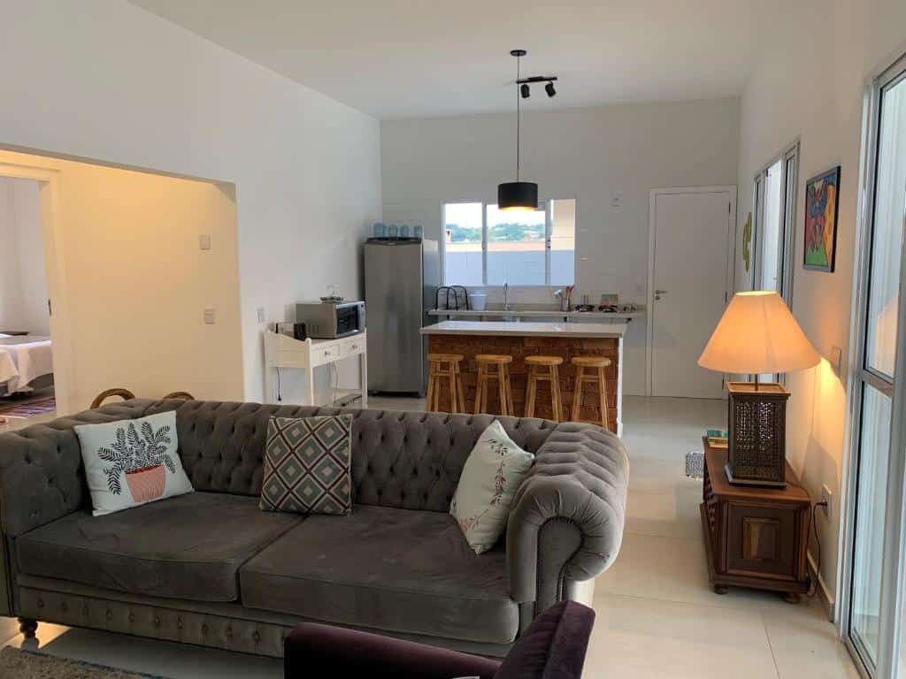 Parte do Casa Charme que mostra o sofá da sala de estar e ao fundo a cozinha da casa com uma bancada com quatro banquinhos, uma pia, uma geladeira e um micro-ondas. No ambiente também tem uma mesinha de madeira com um abajur, um lustre do teto e no canto esquerdo da foto tem uma entrada para outros cômodos. Imagem ilustrando post Airbnb em Brotas.