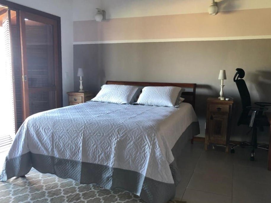 Quarto da Casa com vista para o mar! No meio uma cama de casal, de cada lado uma cômoda com abajur. Do lado direito uma cadeira de trabalho, do lado esquerdo a porta da sacada. Foto para ilustrar post sobre airbnb em Paraty.