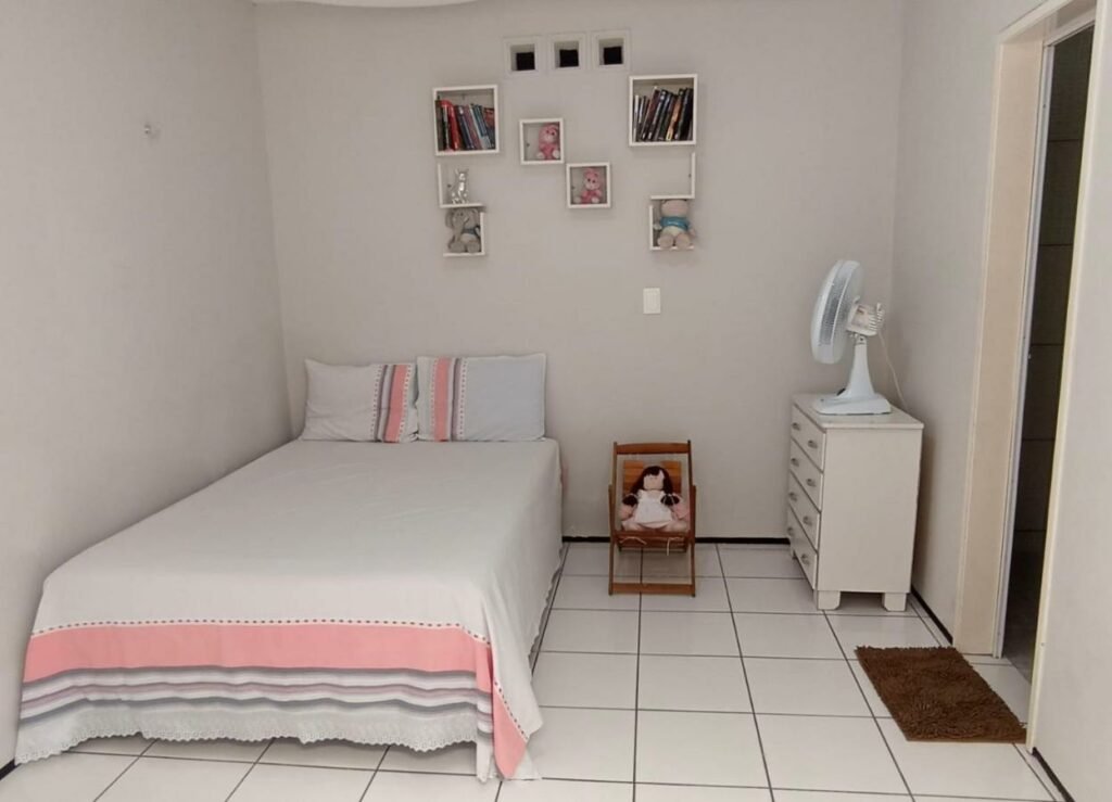 Quarto da Casa da Lena. Uma cama de casal está ao lado esquerdo, ao lado da parede. No meio há uma boneca em um banquinho. Na parede do lado direito está uma cômoda e um ventilador.