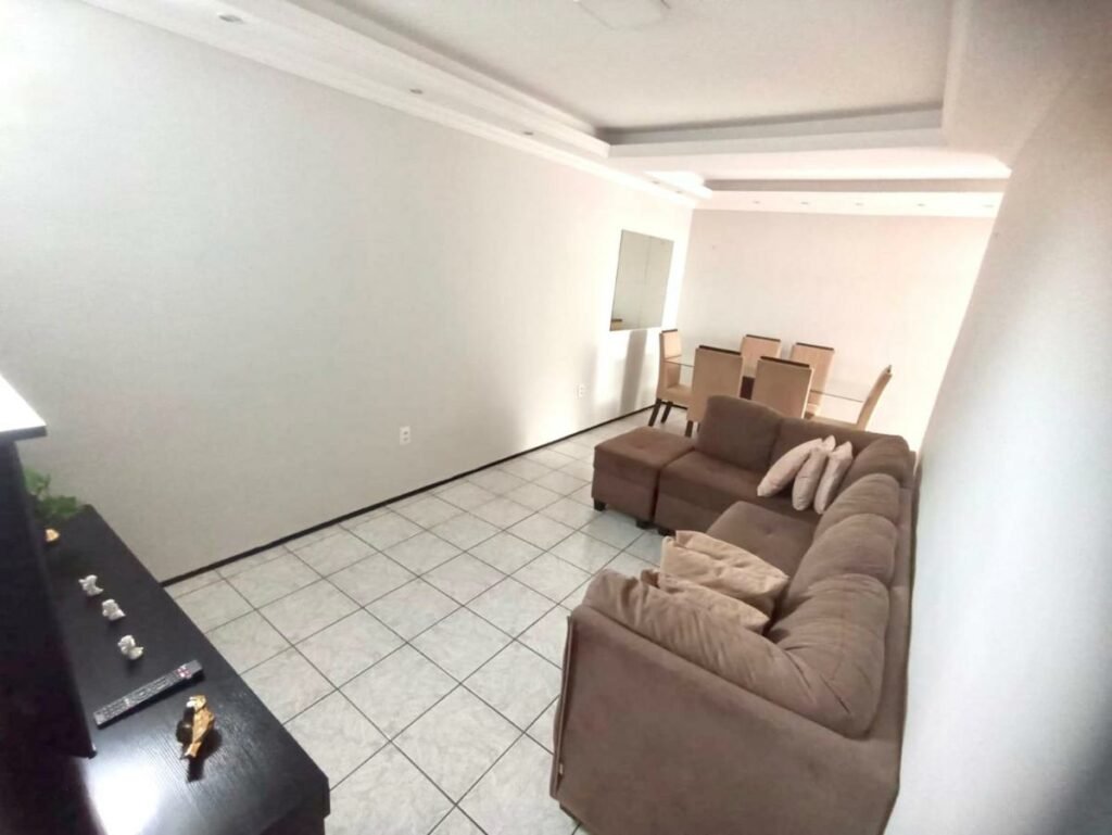 Sala de estar da Casa da Lena. Um sofá de vários lugares está grudado na parede do lado esquerdo. Em sua frente está uma rack com a tv. Ao fundo é possível ver uma mesa de refeições de seis lugares.