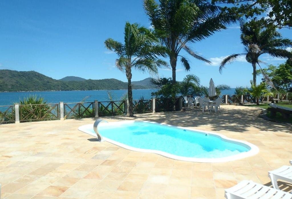 Área externa da Casa frente ao mar c/ piscina. Uma piscina com uma cascata no meio, do lado direito e no fundo cadeiras, jardim e árvores. No fundo a vista da piscina.