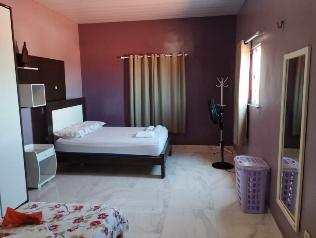 Quarto da Casa inteira em Fortaleza com 02 Quartos sendo 01 Suíte - Praia do Futuro.  Uma cama de casal está ao fundo, perto da parede e ao lado de uma cõmoda. Em sua frente há um ventilador e uma janela. Mais para frente existe outra cama, perto de um espelho pendurado na parede.