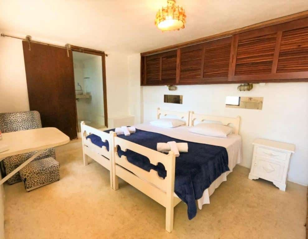 Quarto da Casa Janaina, um dos airbnb em Ilhabela. Duas camas de solteiro com toalhas dobradas aos pés estão encostadas, uma ao lado da outra, na parede ao lado direito. Há mesinhas de cabeceira e espelhos ao lado das duas camas. Em frente há uma mesinha e uma poltrona. Na lateral esquerda das camas há uma porta para o banheiro.