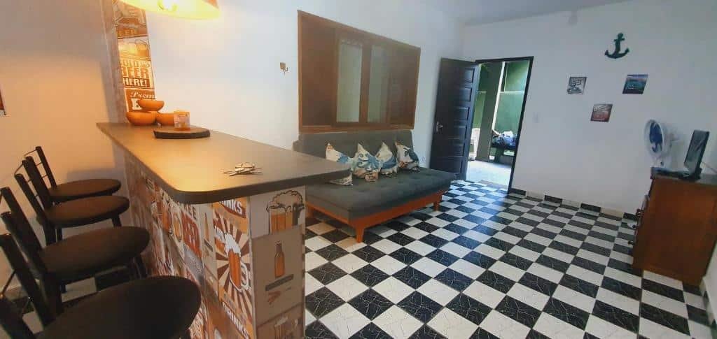 Sala de estar da Casa na Praia da Enseada. Do lado esquerdo uma bancada com banquetas, do lado direito um sofá com uma janela em cima e no fundo a porta, de frente pro sofá uma raque com televisão e um ventilador.