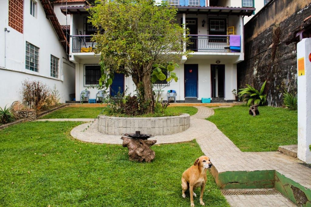 Foto da Casa Pitanga. Nessa foto, um cachorro está parado em um jardim bem cuidado, em frente à pousada de dois andares, criando um ambiente sereno e acolhedor.
