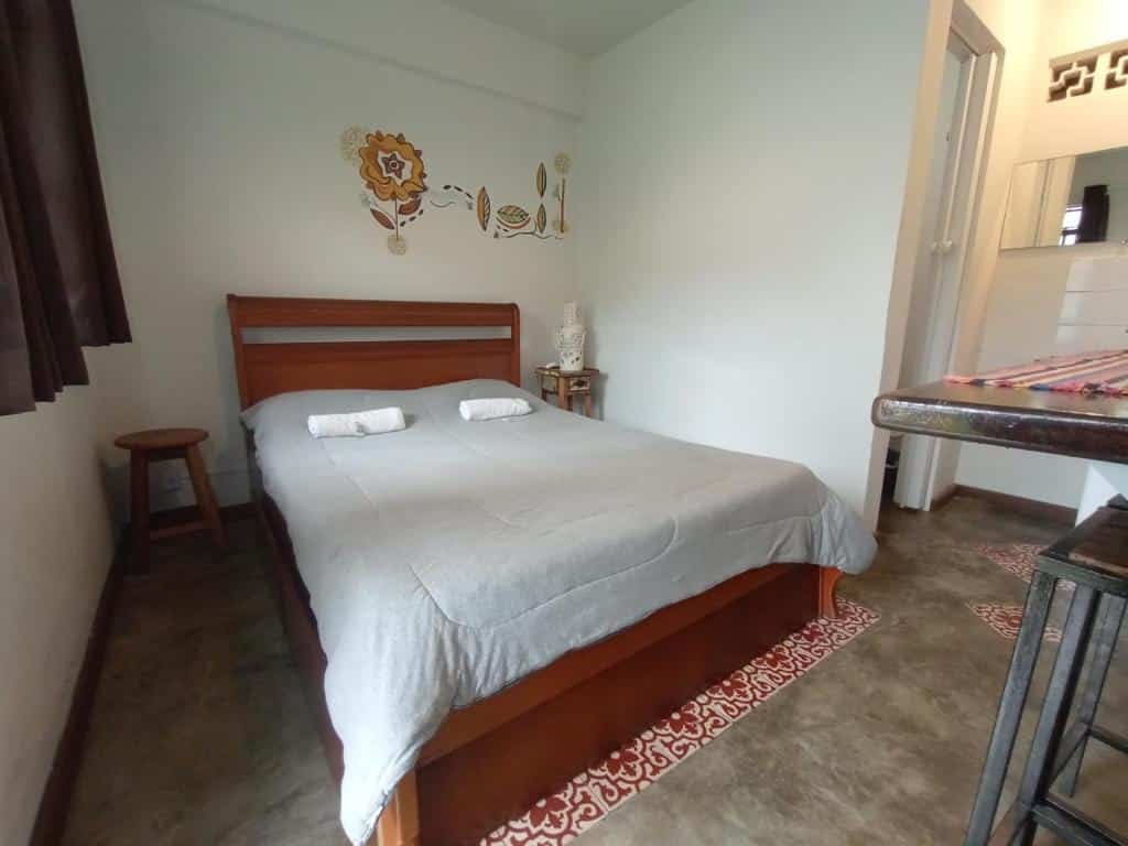 Foto da Casa Pitanga que está ilustrando o post de pousadas pet friendly em Minas Gerais. A foto captura um quarto arrumado com decorações artísticas na parede, oferecendo um vislumbre de um espaço aconchegante e convidativo. A cama de casal está coberta por uma colcha cinza, com dois travesseiros enrolados em cima. Uma decoração de parede estilizada, que parece ser uma flor, fica acima da cama. Ao lado da cama, há um pequeno banco de madeira e um vaso decorativo.