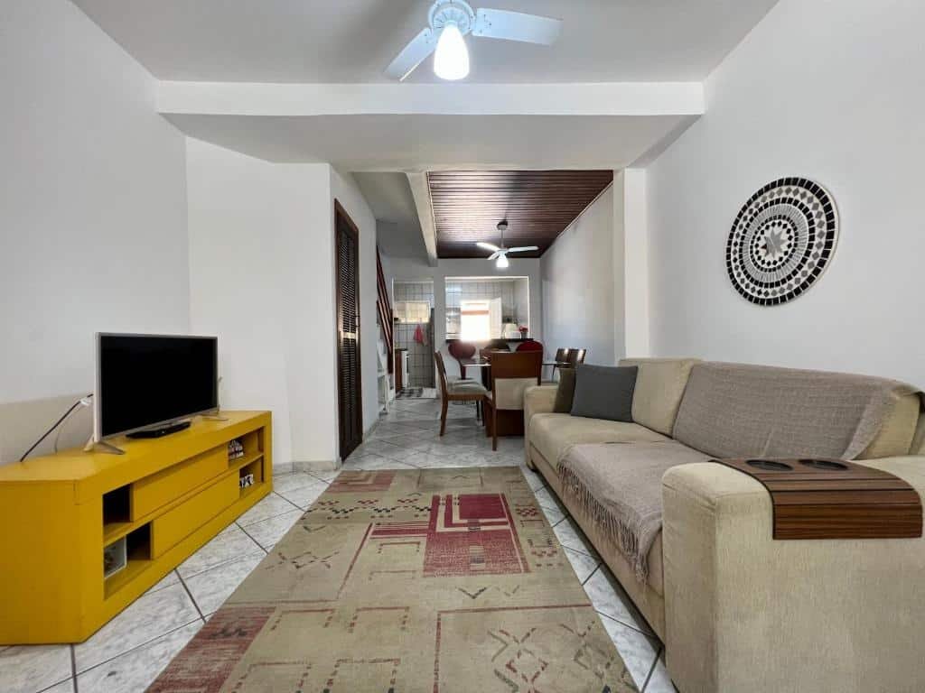 Sala de estar da Casa verde no centro de Paraty. Do lado direito um sofá, de frente uma raque com uma televisão. No fundo, lado direito uma mesa com cadeiras e um ventilador de teto, e a cozinha de fundo.