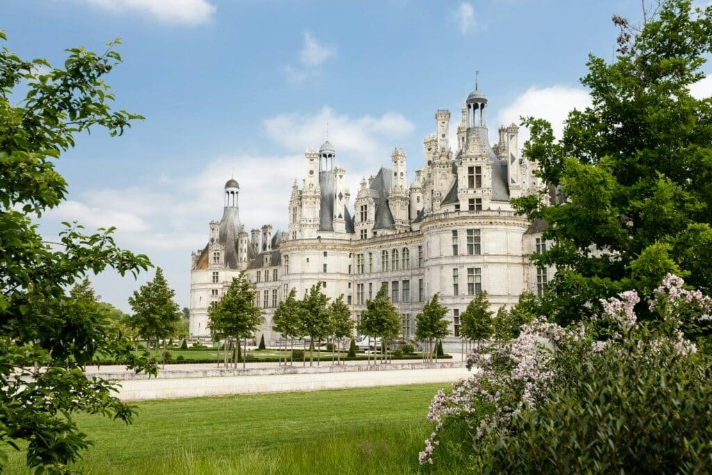 Vista do Château de Chambord, Chambord, França durante o dia com arvores e um belo jardim a frente e ao fundo o castelo.