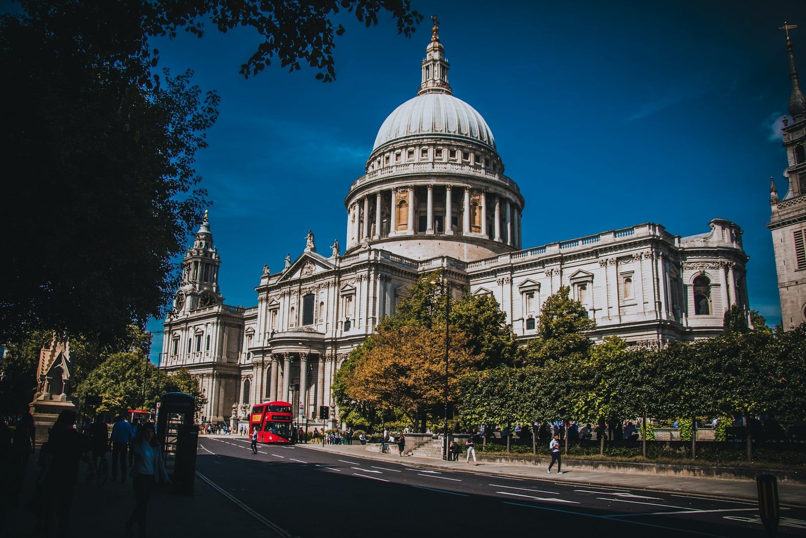Catedral de St. Paul em Londres com sua grande cúpula, cercada por árvores e pessoas. Um ônibus vermelho de dois andares passa na rua.