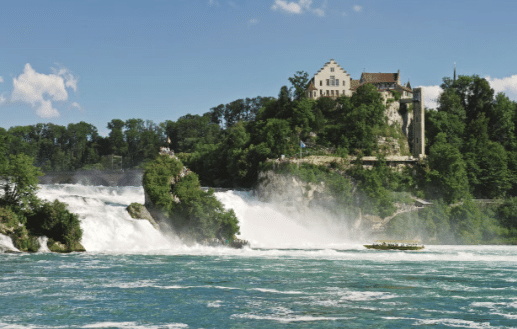 Imagem da Ccataratas do rio Reno grande cachoeira corre cercada por árvores verdes, com um edifício em uma colina e um barco na água sob um céu azul. Representa passeios na Suíça.