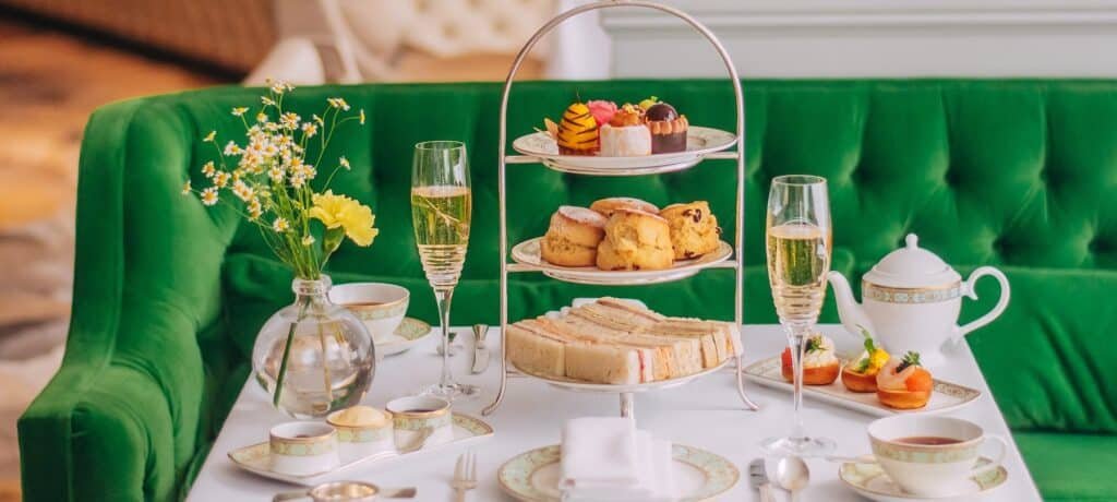 Chá da tarde preparado com uma bandeja em camadas de doces, sanduíches e scones em uma mesa com xícaras de chá, bule e taças de champanhe.