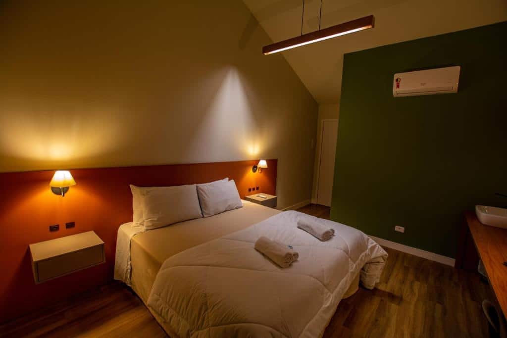 Quarto do Chakras Pousada. Do lado esquerdo uma cama de casal com uma prateleira de cada lado e abajur. Do lado direito um ar-condicionado. Foto para ilustrar post sobre pousadas pet friendly no Paraná.