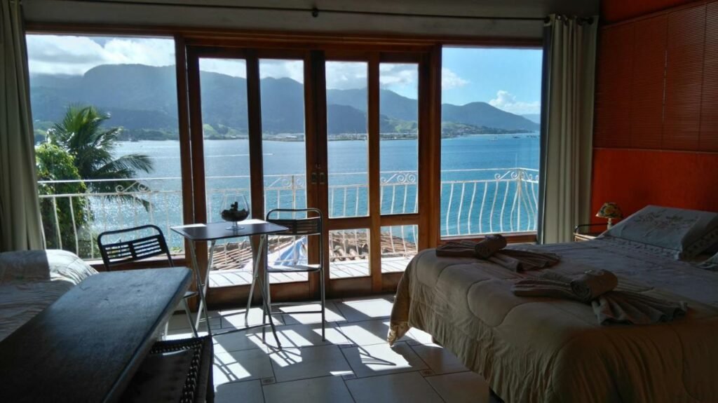 Quarto dos Chalés Bocaina. Uma cama de casal com toalhas aos pés está encostada na parede ao lado direito e encara uma cama de solteiro na parede oposta. Uma mesinha com duas cadeiras fica logo em frente à extensa porta de vidro que leva à varanda com vista para o mar.