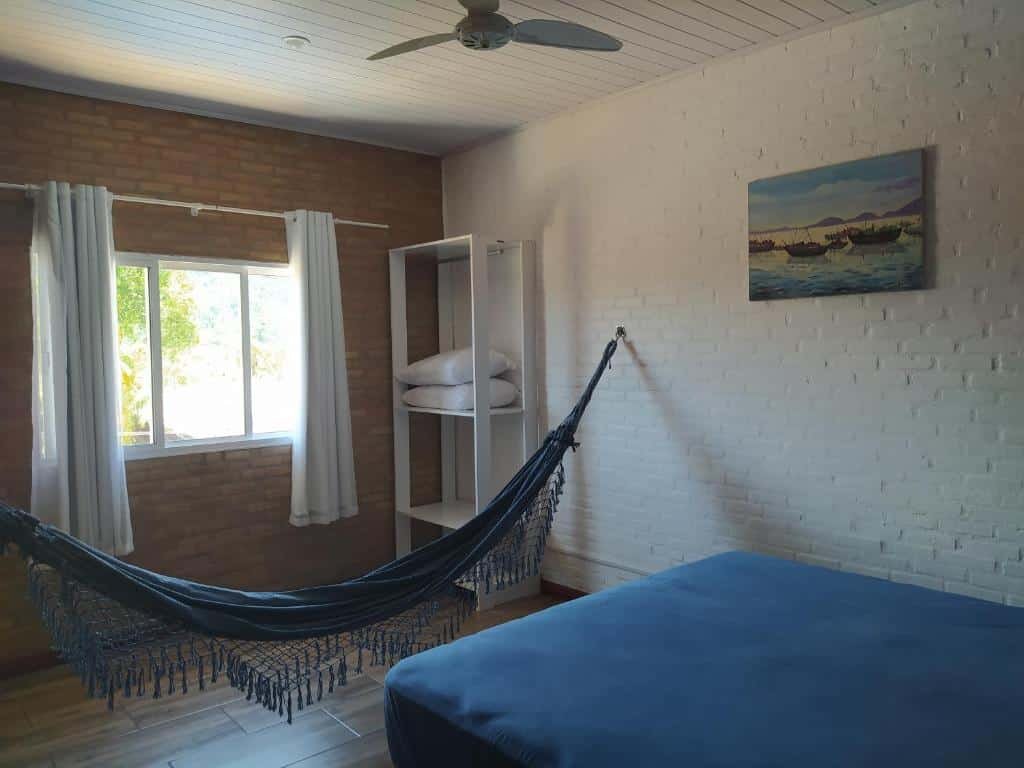 Quarto da Cobertura na Enseada. Uma cama de casal do lado direito, de frente uma rede, uma prateleira e uma janela com cortina.