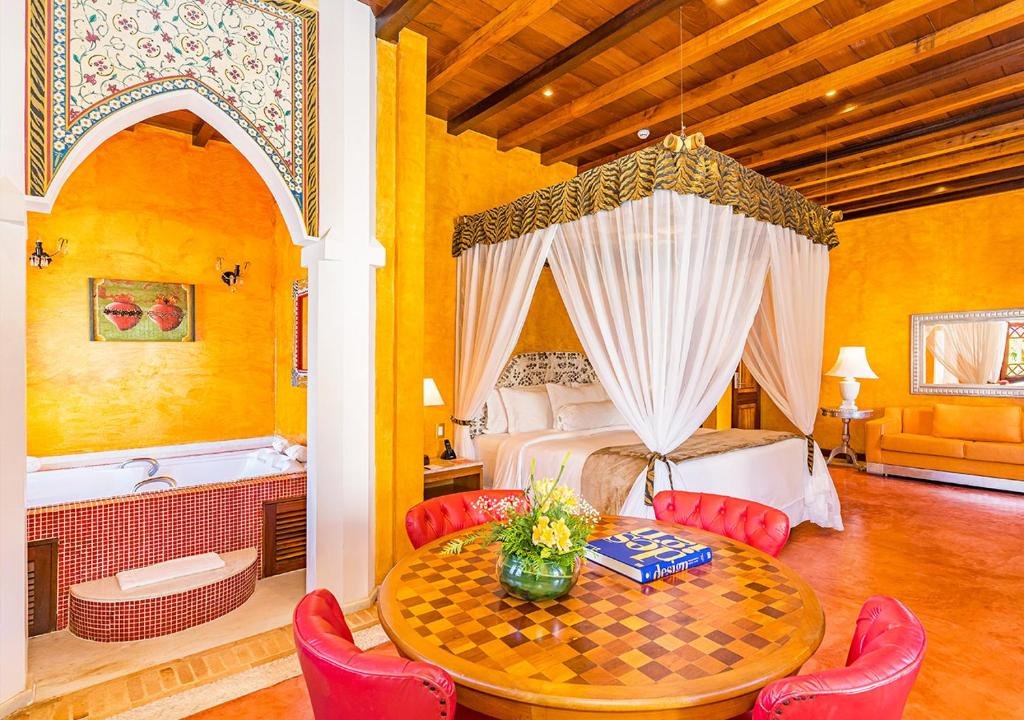 Foto do quarto em DPNY Beach Hotel & SPA Ilhabela. Ilustra o post sobre Onde ficar em Ilhabela. As paredes são laranjas e todos elementos do quarto são coloridos e vivos, em tons de laranja, marrom e vermelho. No primeiro plano há uma mesa com quatro poltronas. Em cima dela há um vaso com flores e um livro. No fundo há uma cama de casal e box, além de um sofá encostado na parede direita. Já na esquerda há uma banheira de hidromassagem.