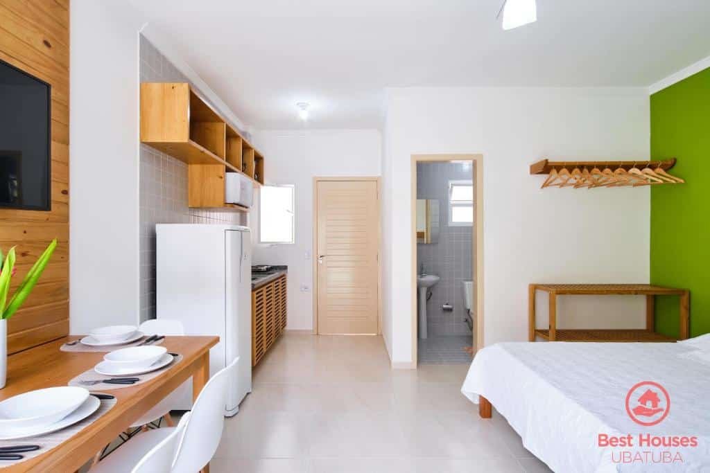 Quarto do Estúdios 3 Praias. Uma cama de casal, uma estante, uma prateleira e uma porta do banheiro do lado direito. Do lado esquerdo uma mesa posta com uma televisão, no fundo uma minicozinha e a porta do quarto. Foto para ilustrar post sobre airbnb na Praia da Enseada.