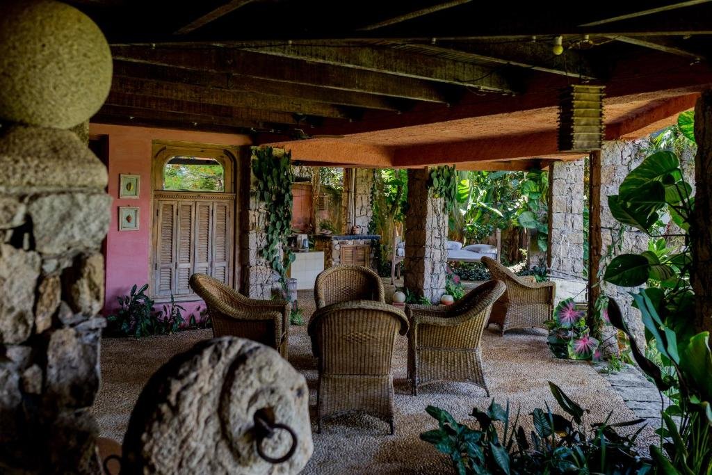Área externa da pousada Feiticeira Guesthouse. Ao centro há quatro grandes cadeiras de palha. Em volta dessas cadeiras há muitas plantas, folhas e pilastras de pedra. Tudo é coberto por um teto de madeira e concreto.