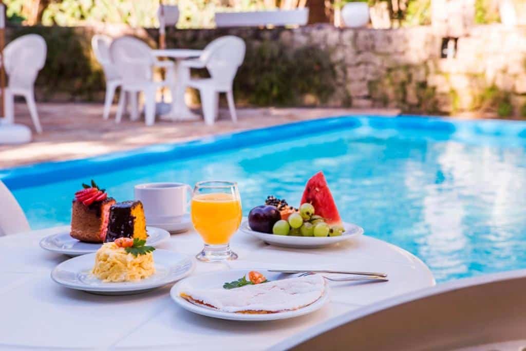 Feiticeira Praia Hotel. Há refeição servida ao lado de uma piscina. A imagem mostra uma mesa com comida e bebida ao lado de uma piscina azul cristalina durante o dia. Há várias opções de comida na mesa, incluindo um bolo de chocolate com cobertura de morango, um prato com ovos mexidos, um prato com frutas frescas e um sanduíche. Uma bebida laranja, possivelmente suco de laranja, está servida em um copo transparente junto a uma xícara branca que pode conter café ou chá. A mesa é branca e está posicionada muito próxima à borda da piscina. Ao fundo, há cadeiras brancas vazias e parte da estrutura do local é visível - parece ser feita de pedras empilhadas ou tijolos. O ambiente é ensolarado e acolhedor, transmitindo uma sensação relaxante.