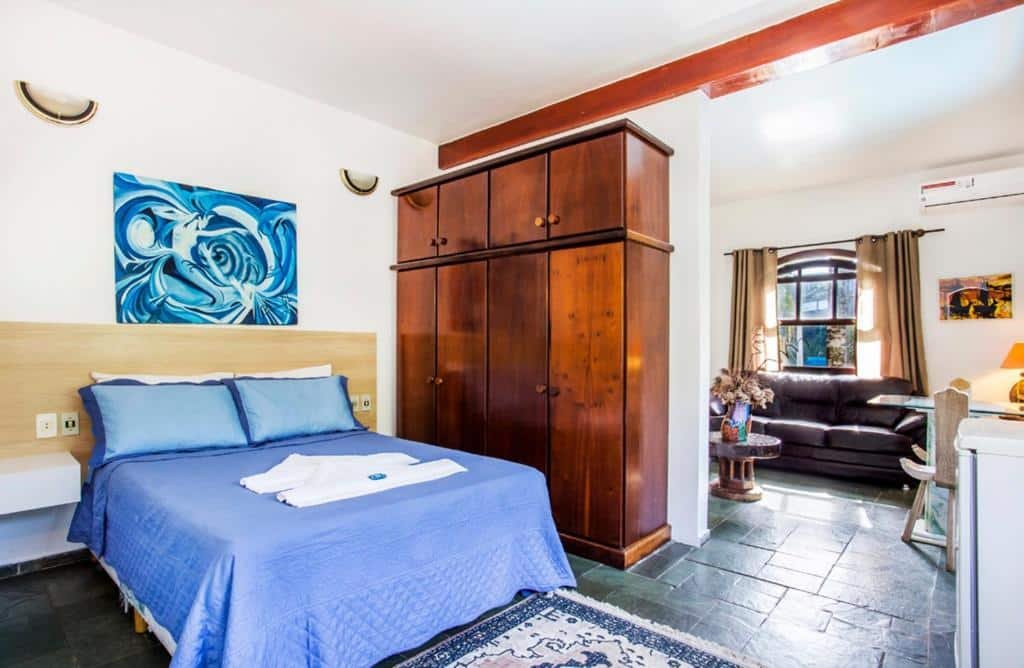 Feiticeira Praia Hotel. Ilustra o post sobre hotéis em Ilhabela. O ambiente retratado é um quarto bem iluminado e aconchegante.
A cama, com lençóis azuis, é o ponto central do quarto. Ela possui uma cabeceira de madeira e está cuidadosamente arrumada.
Uma grande cômoda de madeira escura fica ao lado da cama, oferecendo espaço amplo para guardar pertences.
A janela, coberta por cortinas escuras, permite a entrada de luz natural, iluminando o espaço.
Acima da cama, há uma pintura abstrata em tons de azul, adicionando um toque artístico ao ambiente.
O piso é revestido com azulejos escuros, criando um contraste interessante com os elementos de decoração.
Um sofá preto e macio está posicionado em frente à janela, acompanhado por uma pequena mesa com itens decorativos, como flores.
O teto apresenta vigas de madeira expostas, adicionando um charme rústico a este quarto eclético.
