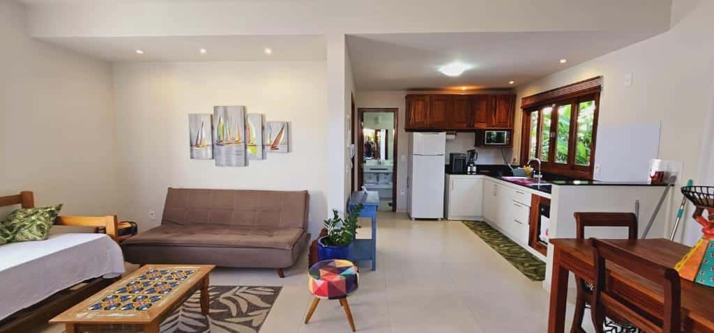 Flat - SERRA E MAR - Paraty. Do lado direito uma mesa de madeira, no fundo uma cozinha com geladeira e microondas, e uma janela em cima da pia. Do lado esquerdo um sofá, um sofá cama, uma mesa de madeira de centro e um banquinho.